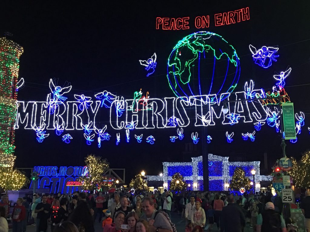 Crowds gather beneath a large illuminated ‘Merry Christmas’ sign, glowing angels, and a neon globe with the words ‘Peace on Earth’ during the Osborne Family Spectacle of Dancing Lights at Disney’s Hollywood Studios.
