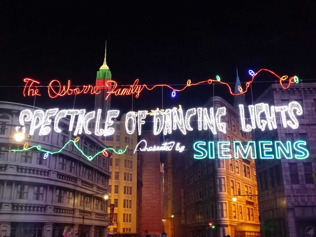 The Osborne Family Spectacle of Dancing Lights sign illuminated in colorful neon letters at Disney’s Hollywood Studios, with holiday light strands and a city street backdrop.