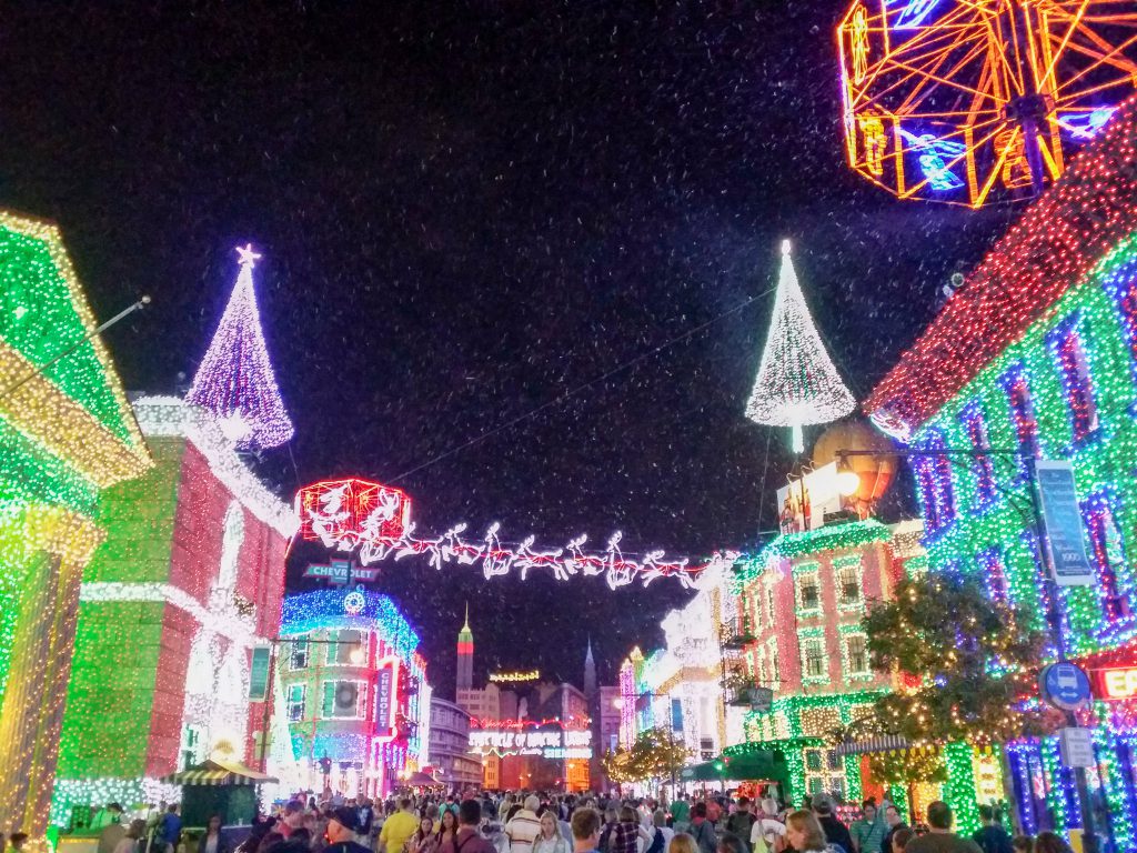 Crowds walking through the Osborne Family Spectacle of Dancing Lights at Disney’s Hollywood Studios, surrounded by colorful buildings covered in Christmas lights, artificial snow, and hanging illuminated Christmas trees.